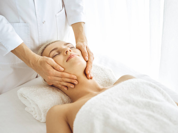 Beautiful blonde woman enjoying facial massage with closed eyes. Relaxing treatment in medicine and spa center concepts - Фото, зображення