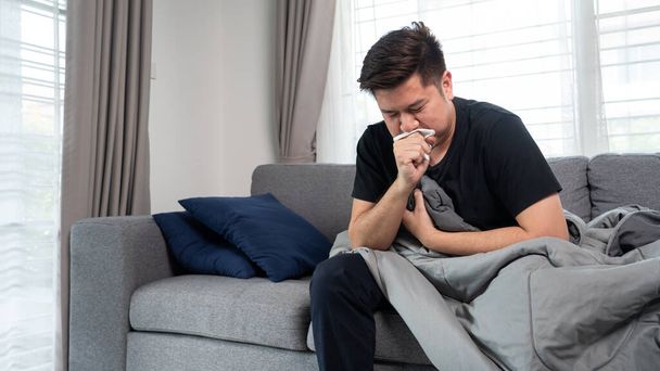 Asiatischer Mann mit saisonalen Infekten Schnupfen und Niesen in Gewebe mit Kopfschmerzen auf Sofa liegend mit hohem Fieber und Grippe. - Foto, Bild