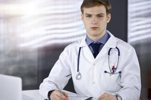 Young male doctor sitting and working with clipboard of medication history record in sunny clinic at his working place. Medicine concept - Foto, Imagem