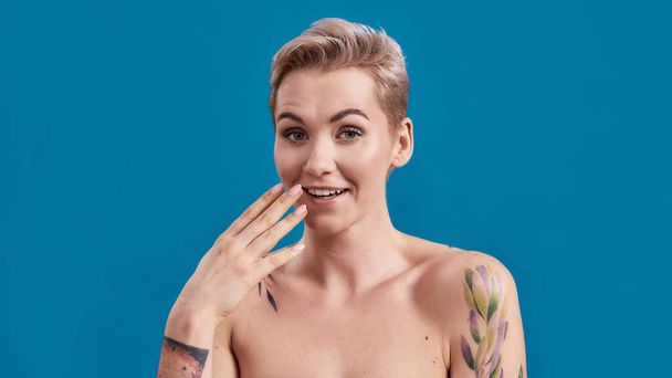 Mocking. Beauty portrait of a young attractive half naked tattooed woman with perfect skin looking surprised at camera with hand near her mouth isolated over blue background - Foto, Imagen