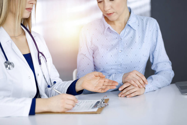 Unknown woman- doctor and her patient are looking at patients current medical test results, while sitting together at the desk in the sunny cabinet in a clinic. Female physician is using a clipboard - Фото, изображение