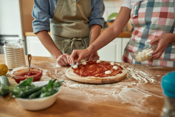 For cheesy taste. Cropped shot of couple making pizza together at home. Man in apron adding, applying tomato sauce on the dough while woman adding mozzarella cheese. Hobby, lifestyle - Фото, изображение