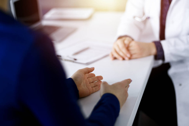 Unknown woman-doctor is talking to her patient about her diagnosis, while sitting together at the desk in the sunny cabinet in a clinic. Physician at working place, close-up. Perfect medical service - Foto, imagen