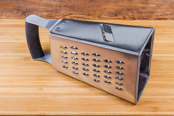Stainless steel kitchen box-shaped grater with four various grating and slicing surfaces lies on the wooden cutting board with vegetables slicer up - Fotografie, Obrázek