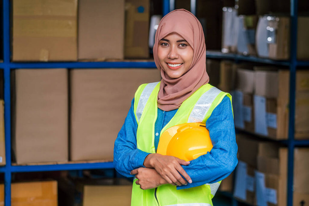 Porträt einer muslimischen Arbeiterin, die Hijab trägt und im örtlichen Lager steht und einen Helm hält, bevor sie ihre Arbeit aufnimmt, Geschäft und Industrie, Wiedereröffnung des Geschäfts nach Covid19-Ausbruch - Foto, Bild