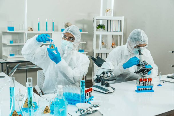 Δύο επιστήμονες με κοστούμι PPE εργάζονται στο εργαστήριο, ενώ τον έλεγχο των αποτελεσμάτων των εξετάσεων του δείγματος αίματος. Μπλε τόνος. SARS-CoV-2, Covid-19 ΘΕΜΑ. - Φωτογραφία, εικόνα