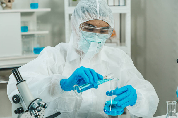 Hombre científico en traje PPE que trabaja en laboratorio mientras se vierte la solución de cilindro a Erlenmeyer matraz con signo de peligro biológico. Tono azul. SARS-CoV-2, Covid-19 TEMA. - Foto, imagen