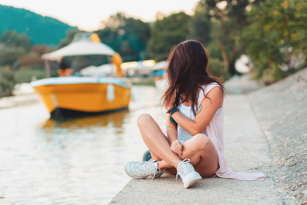 Une femme s'assoit les jambes croisées sur une jetée au bord de la rivière, regardant un bateau qui passe. Le concept des vacances d'été. - Photo, image