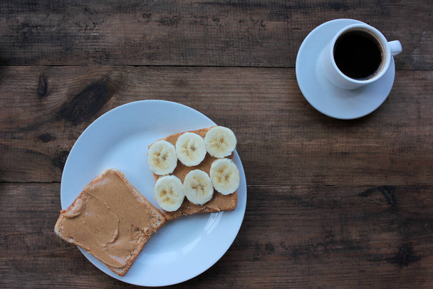 Cup of coffee and peanut butter banana toast on wooden background. Slices of whole wheat bran bread with peanut paste on a plate. Healthy breakfast - Zdjęcie, obraz