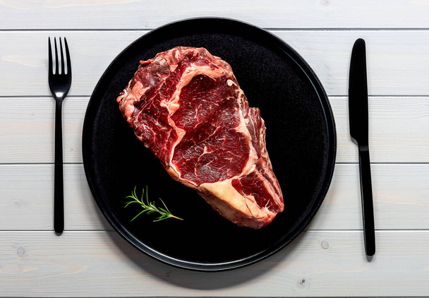 Raw beef rib eye steak. Organic farm meat. Black plate on wooden table - 写真・画像
