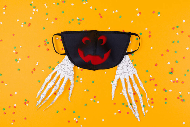Újrafelhasználható fekete antivírus maszk Jack Lámpás lenyomatokkal és papírcsontváz kezekkel. Narancs háttér dekoratív csillagok. Lapos fekvés. A vírus elleni védelem fogalma Halloweenkor. - Fotó, kép