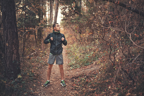 Pojetí sportu a aktivního životního stylu. Mladý muž v černé teplákové soupravě s batohem na zádech, pózující v podzimním lese. Tmavé barvy. - Fotografie, Obrázek