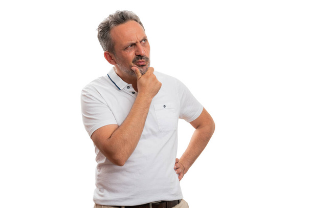 Homme adulte avec une expression curieuse pensant toucher le menton en utilisant la main portant un t-shirt d'été décontracté regardant la zone d'copyspace vierge pour le texte publicitaire isolé sur fond de studio blanc - Photo, image