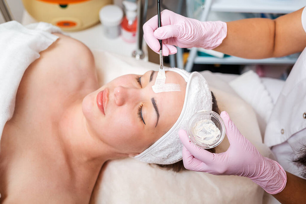 The cosmetologist in medical gloves holds a bowl with a mask, applying cosmetics with a brush on the client's face. Top view. Concept of professional cosmetology and treatment. - Photo, Image