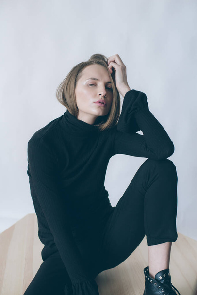 Young attractive girl model with blond hair and nude makeup in black knee socks and black trousers posing in the studio on a white background sitting on the table - Foto, afbeelding