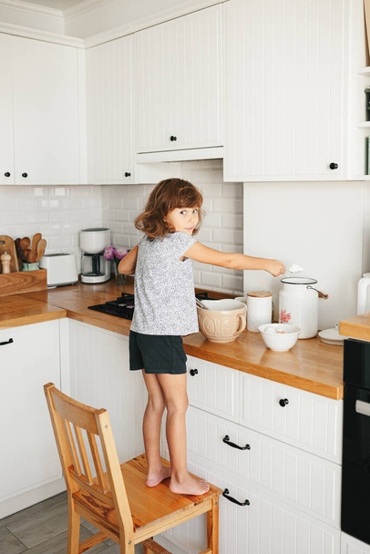 Child girl preparing dough for pancakes at the kitchen. Concept of food preparation, white kitchen on background. Casual lifestyle photo series in real life interior - Zdjęcie, obraz