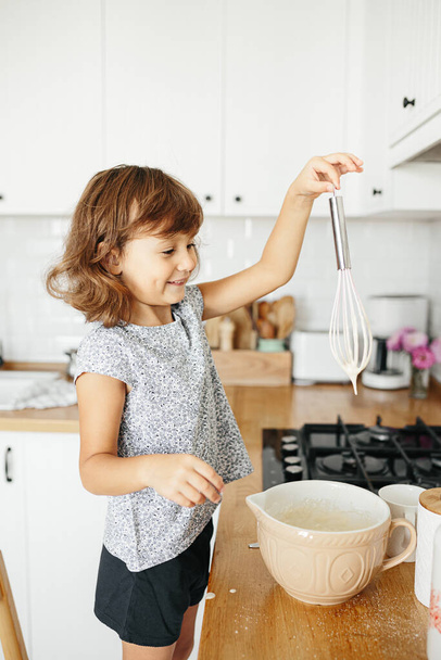Child girl preparing dough for pancakes at the kitchen. Concept of food preparation, white kitchen on background. Casual lifestyle photo series in real life interior - Foto, Imagem