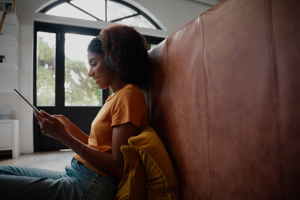 Side view of young african woman sitting on floor using digital tablet with headphones while on video call - Фото, зображення