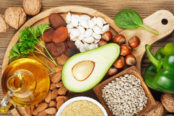 Foods rich in vitamin E such as wheat germ oil, dried wheat germ, dried apricots, hazelnuts, almonds, parsley leaves, avocado, walnuts, pumpkin seeds, sunflower seeds, spinach and green paprika on wooden table - Valokuva, kuva
