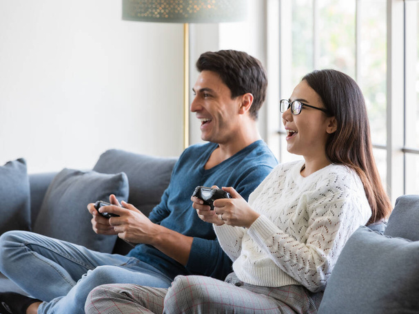 Yakışıklı beyaz adam ve güzel Asyalı kadın aşığı evde oturma odasında oturuyor ve birlikte oyun oynamak için joystick kullanıyorlar mutluluk ve neşeyle. Çeşitli çiftler ve ev konsepti.. - Fotoğraf, Görsel