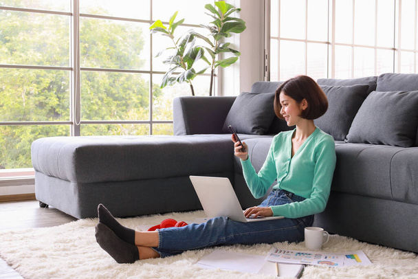 Young and beautiful Asian woman sitting in house beside sofa with easy and cozy manner using laptop notebook computer to work and study in private area. Idea for working and learning from home. - Фото, изображение
