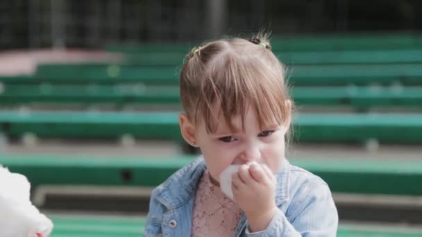 Kaunis ja onnellinen pieni tyttö syö hattaraa penkillä. - Materiaali, video