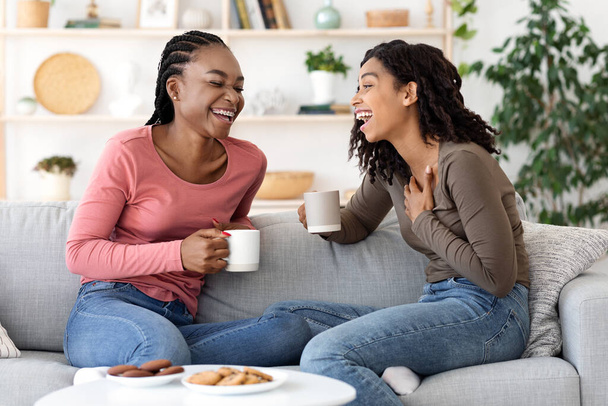 Two african besties laughing while drinking tea together at home - Photo, Image