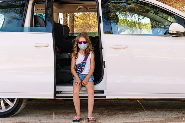 Dívka s maskou na tváři a růžové sluneční brýle sedí na dveřích v autě s otevřenými dveřmi, zatímco ona jde na dovolenou uprostřed covid19 coronavirus pandemie - Fotografie, Obrázek