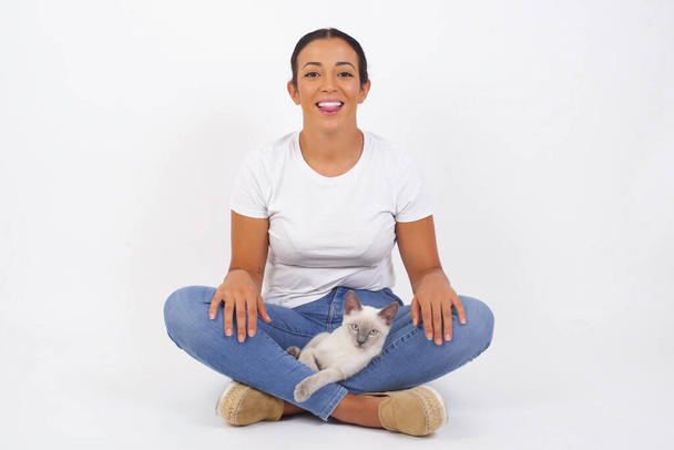 Beautiful Young woman with happy and funny face smiling and showing tongue. Wearing casual clothes and standing against gray studio background. Beautiful Arab woman with cute cat  - Zdjęcie, obraz