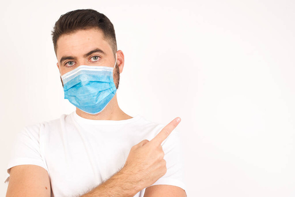 Excited surprised man wearing medical mask points happily aside on copy space of gray background for your fashion clothing advertising information or promotional text. - Fotografie, Obrázek