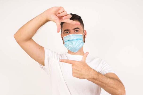Portrait of young positive man wearing medical mask with cheerful expression, has good mood, gestures finger frame actively at camera. - Photo, Image