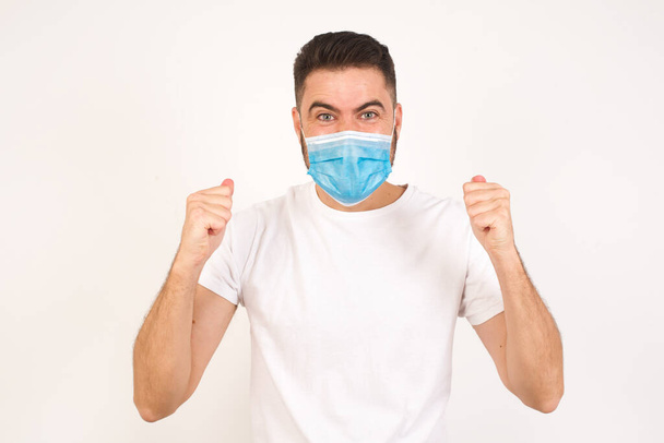Waist up portrait of strong successful determined young male winner in casual t-shirt raising arms, clenching fists, exclaiming with joy and excitement. Victory, success and achievement concept. Caucasian man wearing medical mask  - Photo, Image