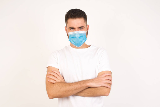 Picture of angry young handsome caucasian man wearing medical mask crossing arms standing isolated over white background. Looking at camera with disappointed expression. - Zdjęcie, obraz