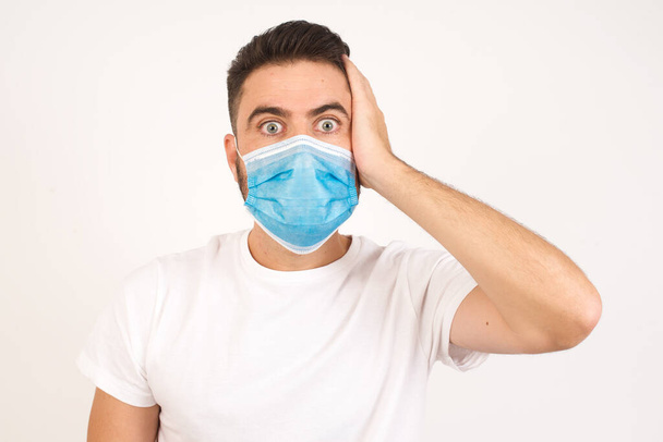 Worried and anxious caucasian man wearing medical mask can not remember anything. Portrait of confused cute boy wearing casual clothes, holding hand on hair and frowning, panicking, losing memory over white wall - 写真・画像