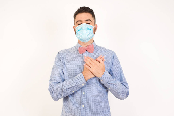 Faithful man wearing medical mask closes eyes and keeps hands on chest near heart, shows his kindness or favour, expresses sincere emotions, being kind hearted and honest. Body language and real feelings concept - Photo, Image