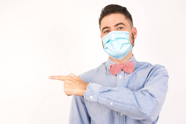 Look over there! Handsome blond caucasian man wearing medical mask pointing away and smiling to you while standing against grey background. - Photo, Image