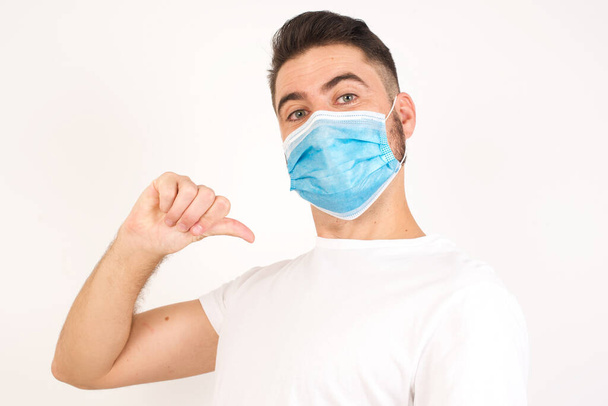 Closeup of cheerful handsome European dark haired blue eyed man looks joyful, satisfied and confident, points at himself with thumb, isolated over gray studio background. Caucasian man wearing medical mask - Foto, Imagem
