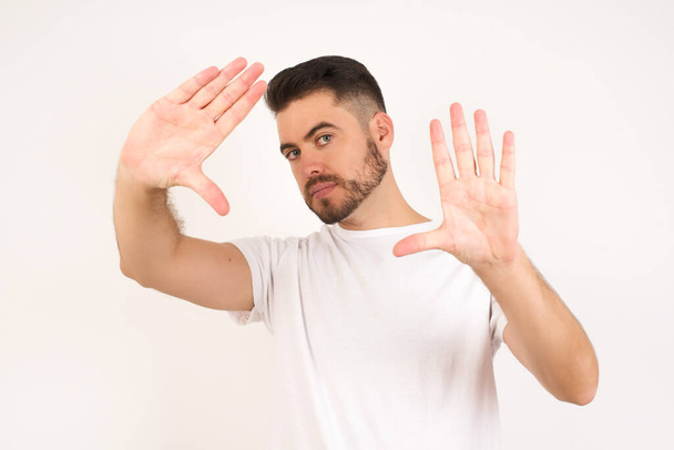 Gesturing finger frame. Portrait of smiling young caucasian man wearing white t-shirt looking at camera and gesturing finger frame while standing against gray studio wall. - Foto, imagen