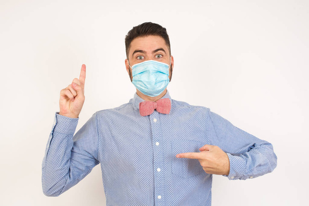 Positive glad male says: wow how exciting it is, has amazed expression, indicates something. Joyful man wearing medical mask demonstrates something. One hand on her chest and pointing with other hand. - 写真・画像