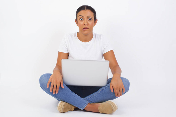 Beautiful young woman with laptop nervous and scared biting lips looking camera with impatient expression, pensive  - 写真・画像