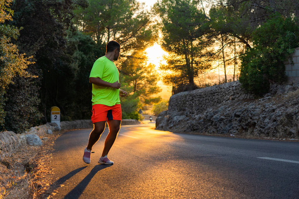 jeune homme espagnol courant sur une route dans la forêt de pins au coucher du soleil regardant en arrière, portant des vêtements de sport flashy - Photo, image
