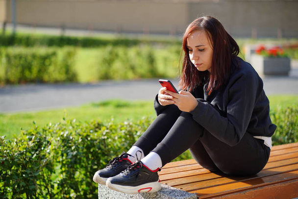 Young woman sitting on a Park bench, looking at her mobile phone. A woman using a smartphone uses the Internet in the courtyard of a residential building - 写真・画像