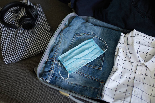 Packing suitcase at home with casual man items, stuff nad protection mask during pandemic coronavirus for travel, journey, vacation . Copy space. - 写真・画像