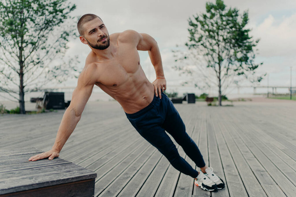 Young handsome shirtless ordinary bearded man stands in diagonal body position, makes side plank, practices endurance and stays in good physical shape. Sportsman in active wear trains outdoor - Photo, Image