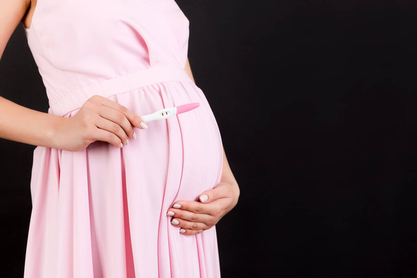 Imagen recortada de la mujer embarazada en vestido rosa que muestra prueba de embarazo positiva con dos rayas en el fondo negro. Embarazo saludable. Copiar espacio. - Foto, Imagen