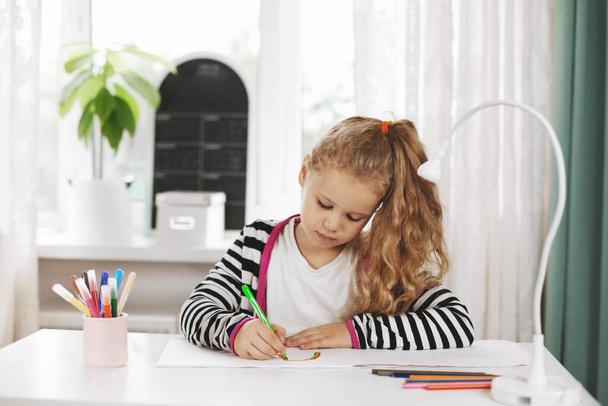 Uma garota encantadora se senta em uma mesa e desenha um arco-íris em um álbum com lápis coloridos. Trabalho de casa, formação em casa, distância social - Foto, Imagem