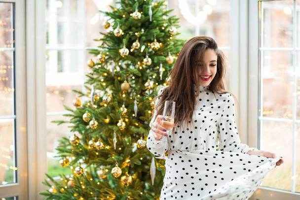Alegre joven hembra con copa de champán de pie contra el brillante árbol de Navidad iluminado - Foto, imagen