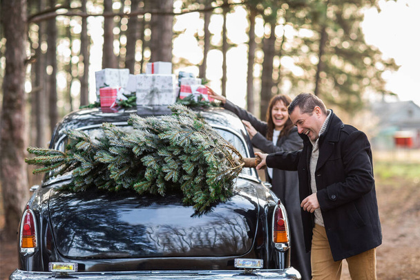 Homem amoroso e mulher pelo carro retro com presentes de Natal no telhado e árvore para celebração no parque - Foto, Imagem