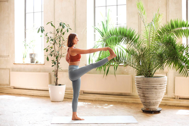 Adatta donna adulta seduta su un materassino in posa e distogliendo lo sguardo mentre pratichi yoga in una spaziosa stanza a casa - Foto, immagini