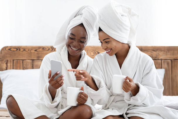 Onnelliset mustat naiset kylpytakit juovat kahvia älypuhelimella - Valokuva, kuva
